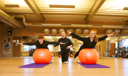 Ein junger Mann und eine junge Frau führen eine Übung auf einem Gymnastikball unter Aufsicht eines Trainers durch