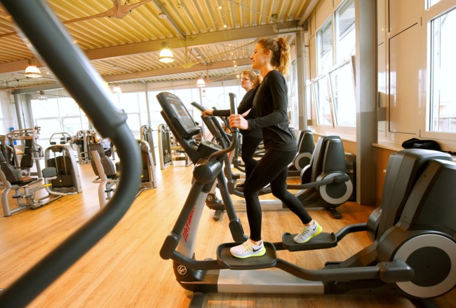 Foto: Zwei Damen trainieren auf einem Cross-Trainer im Aktiv-Fitnessstudio, im Hintergrund befinden sich weitere Fitnerss-Geräte
